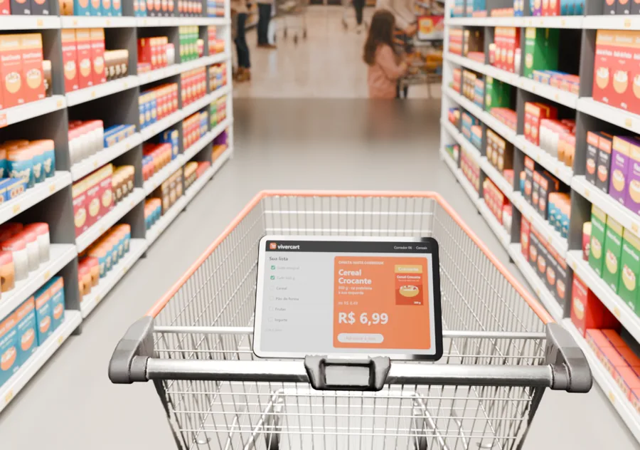 Carrinho de supermercado comum com a tela ViverCart instalada na alça, mostrando a oferta do corredor de cereais